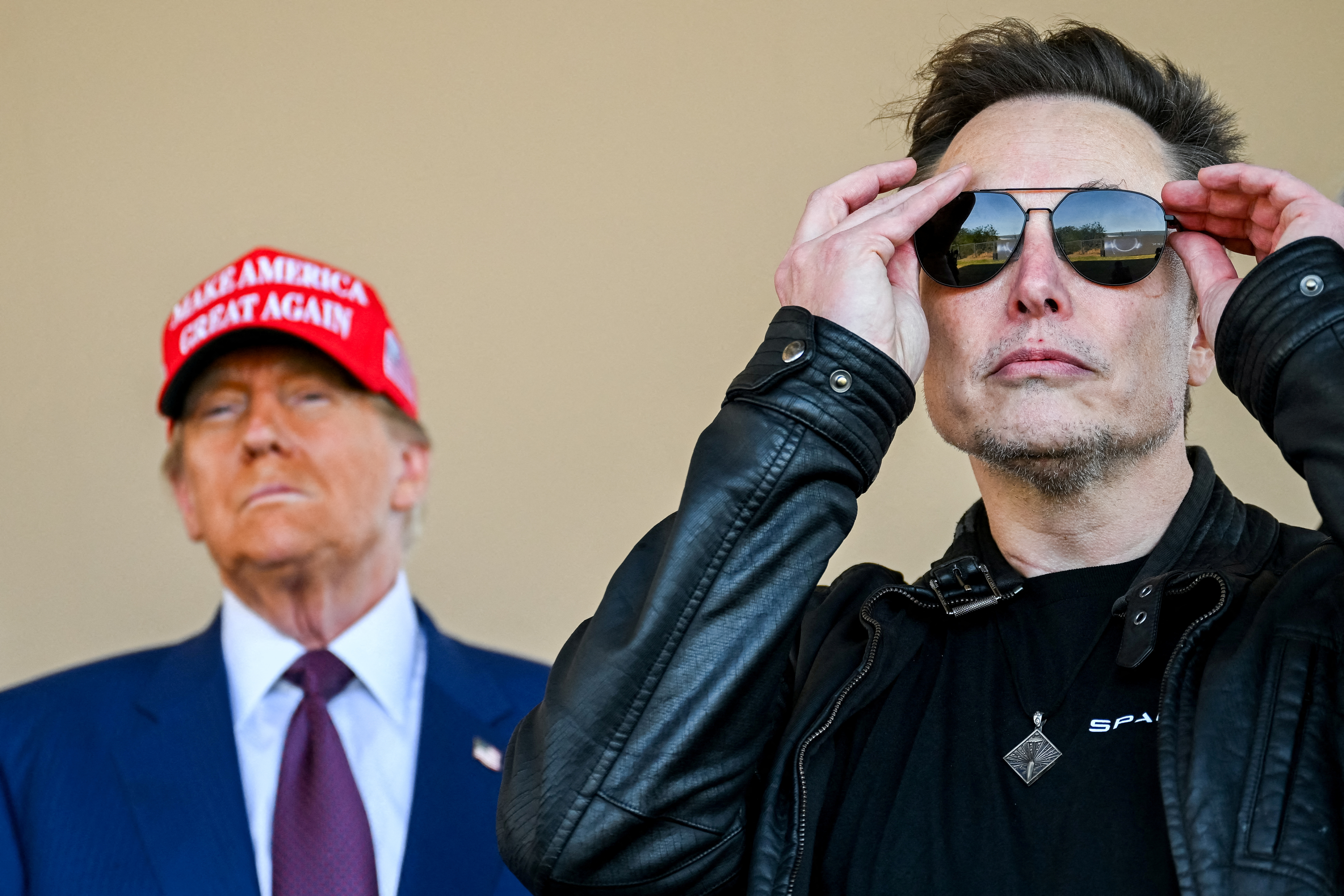 Elon Musk adjusting his sunglasses with Donald Trump in the background wearing a "Make America Great Again" hat.