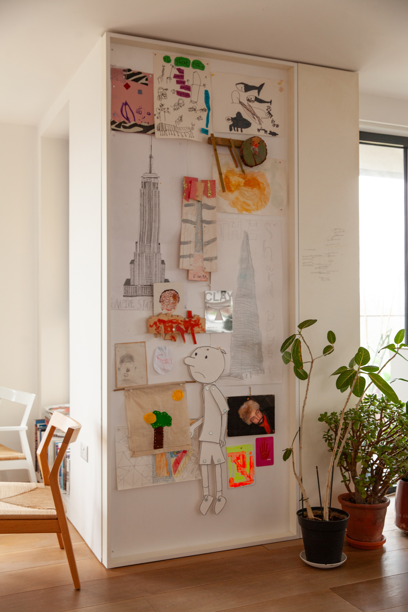 A white wall covered in children's drawings and artwork, next to potted plants and a wooden chair.