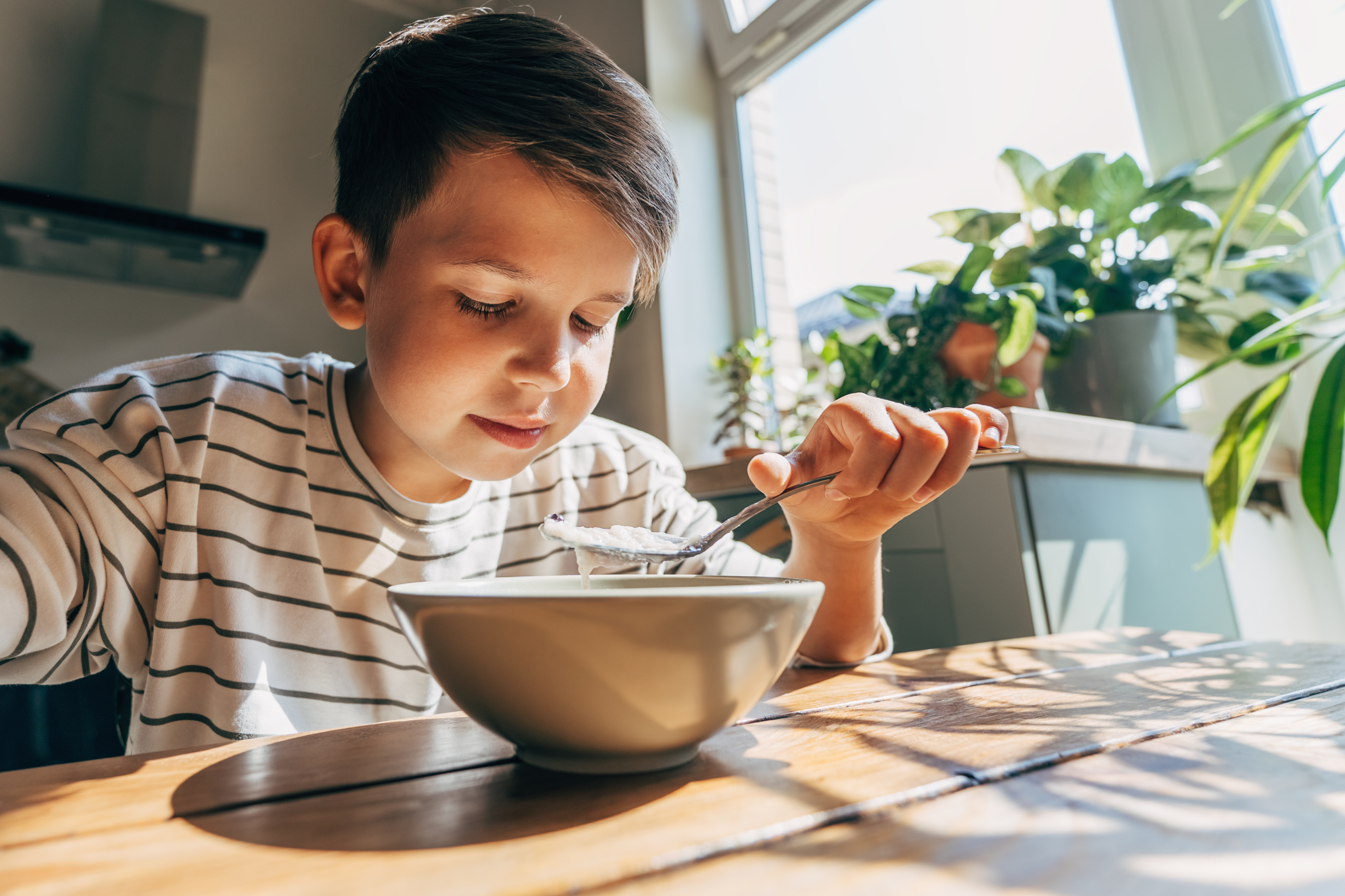 A boy eating breakfast porridge with a spoon by a window.