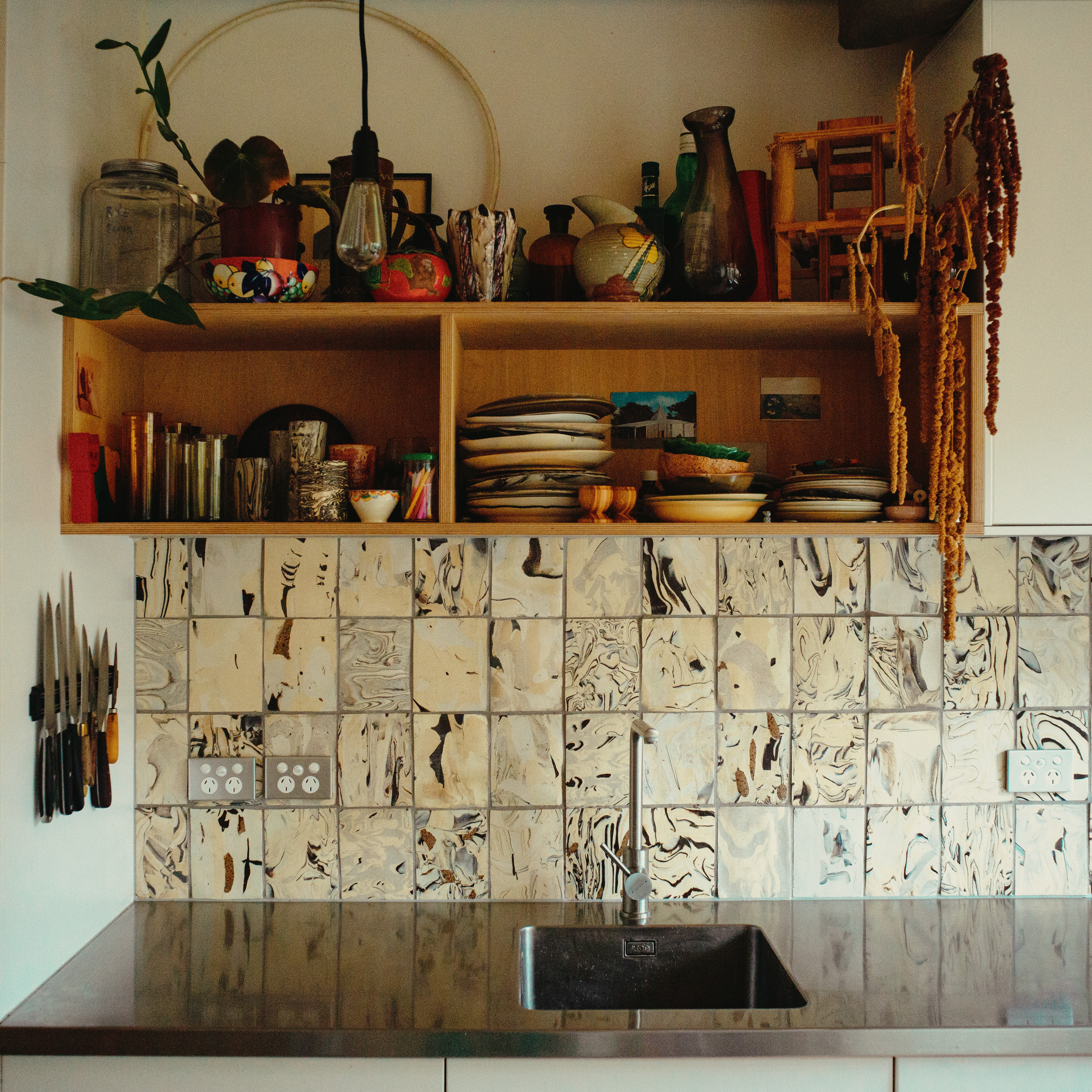 A kitchen with a stainless steel counter, a square sink, white and beige marbled tiles on the wall, and wooden shelves above holding dishes and decorative items.