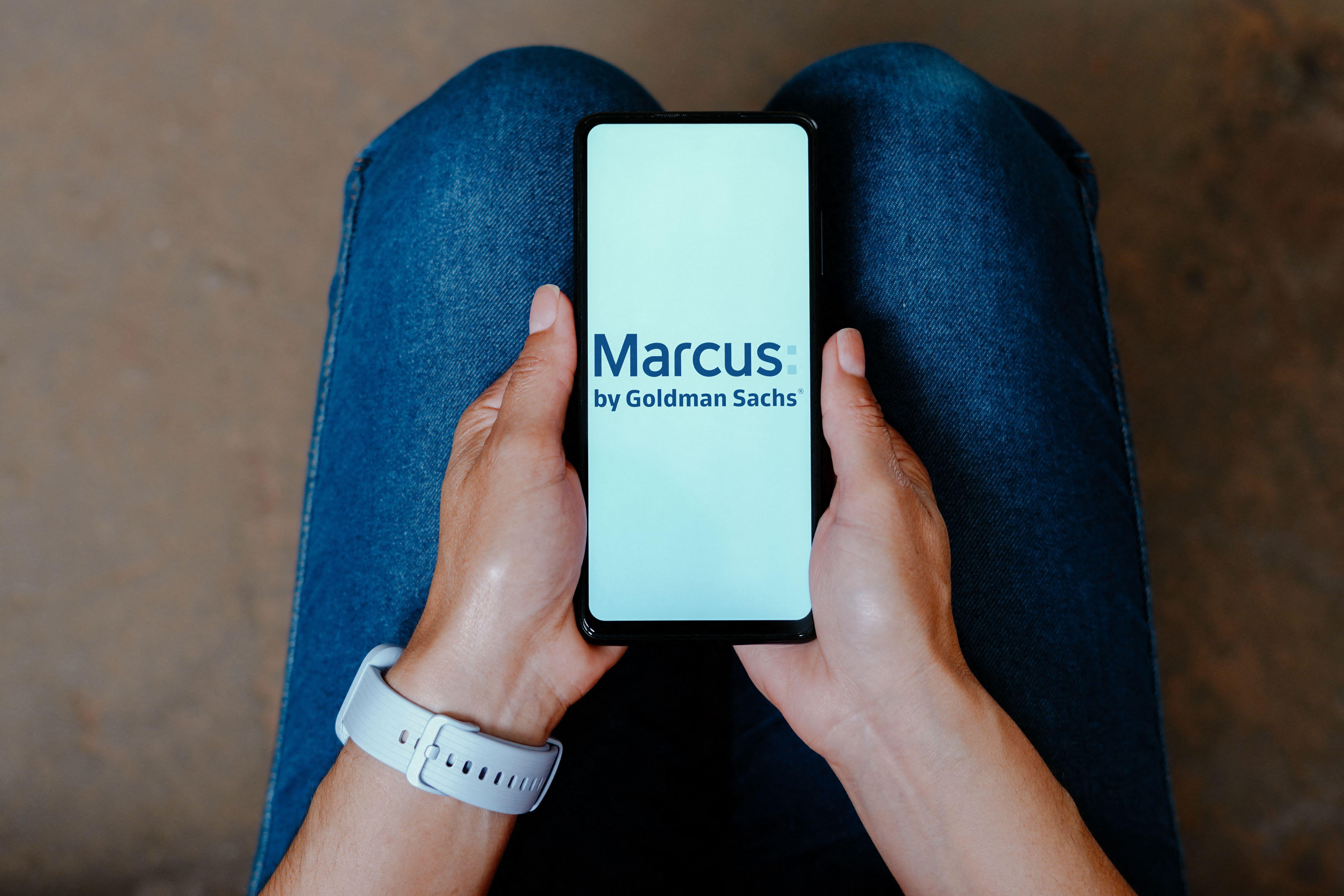 Illustration of the Marcus by Goldman Sachs logo on a smartphone held by a person in blue jeans.