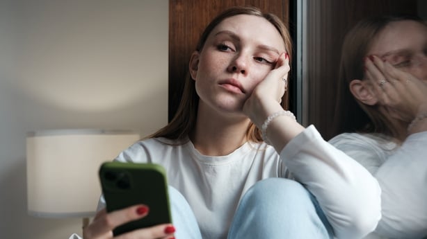  Upset redhead teen girl sitting by window with phone waiting call from boyfriend, feeling sad and depressed woman using smartphone. Social Media depression in teens