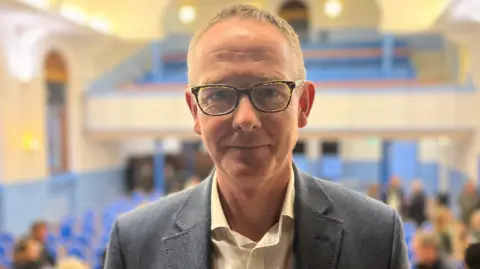 A man with short hair and glasses looks at the camera