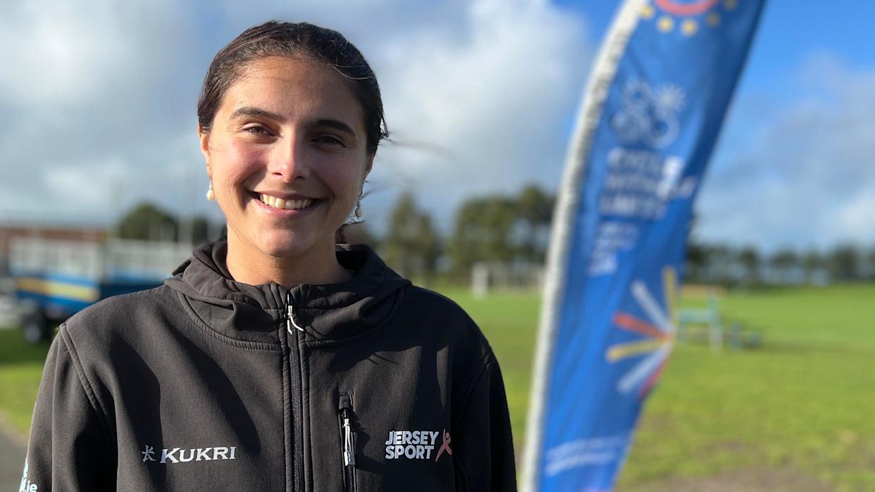 Claudia is wearing a black Jersey Sport Kukri jacket and stands outdoors beside a grassy sports field, with a blue event flag in the background under a partly cloudy sky.