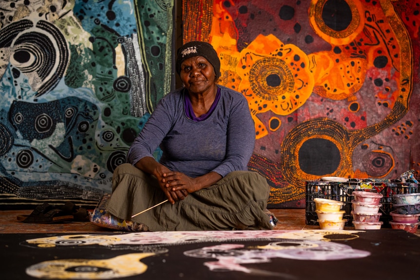 A woman wearing a beanie, a purple top and a green / grey skirt sits on the floor with a big canvas in front and two on the wall