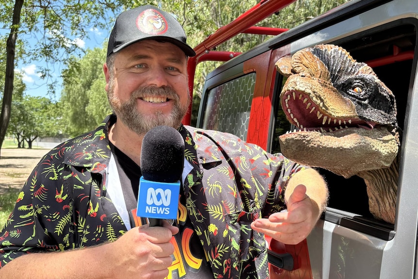 A man in colourful shirt holds an ABC microphone, one arm leans on a car window where a model dinosaur pokes its head out