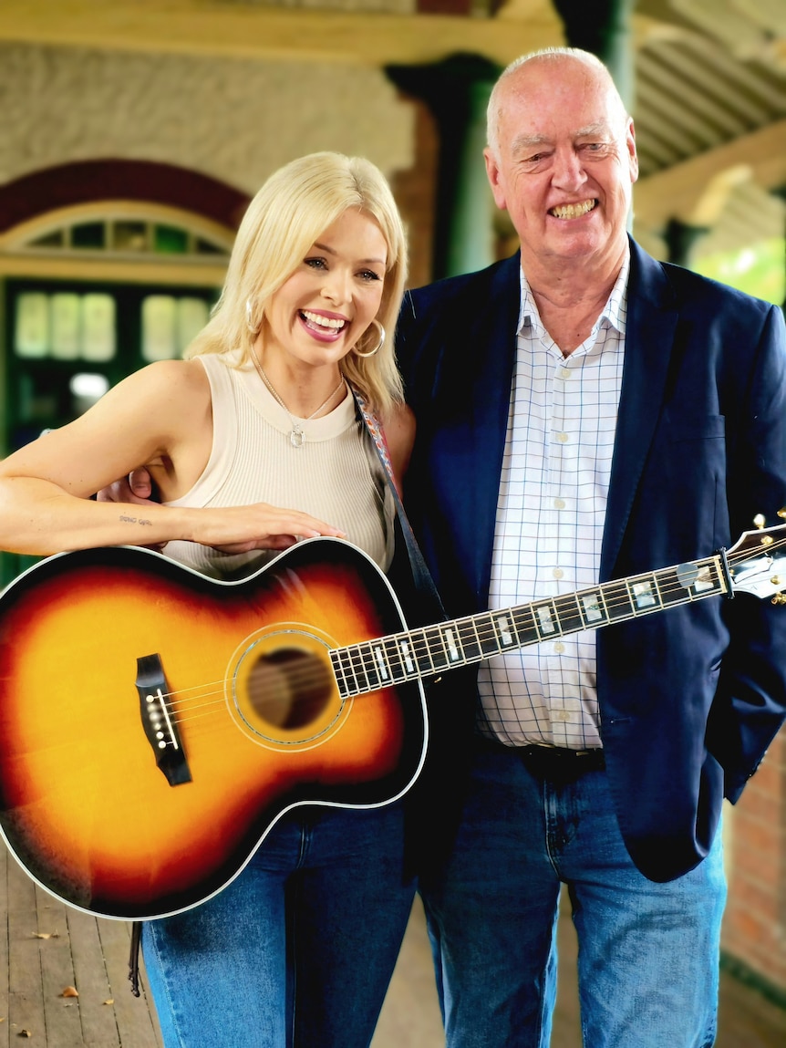 A blonde woman in a singlet and guitar standing alongside a bald man in a blue blazer