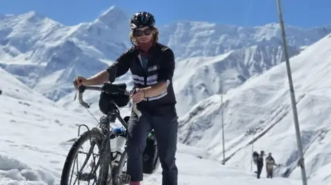 Olly Hargreaves Olly pushes his bike through snow-covered mountains. He wears a black bike helmet, a black cycling top and dark grey trousers.