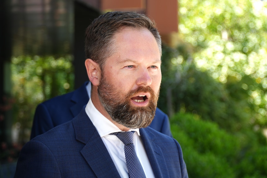 Adam Hort, wearing a suit and tie, speaks at a press conference.