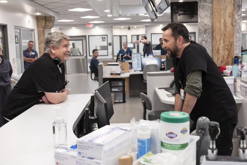 John Wells, 69, headphones around his neck, and Noah Wyle, 54, look at each other over a desk in a hospital ER set.