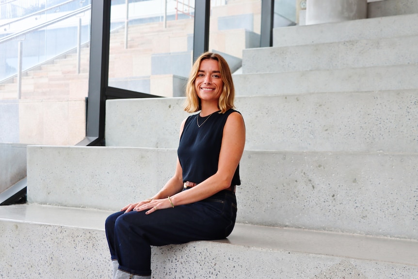 A smiling young woman, Alexis Hutcheon, sits on a stairwell.
