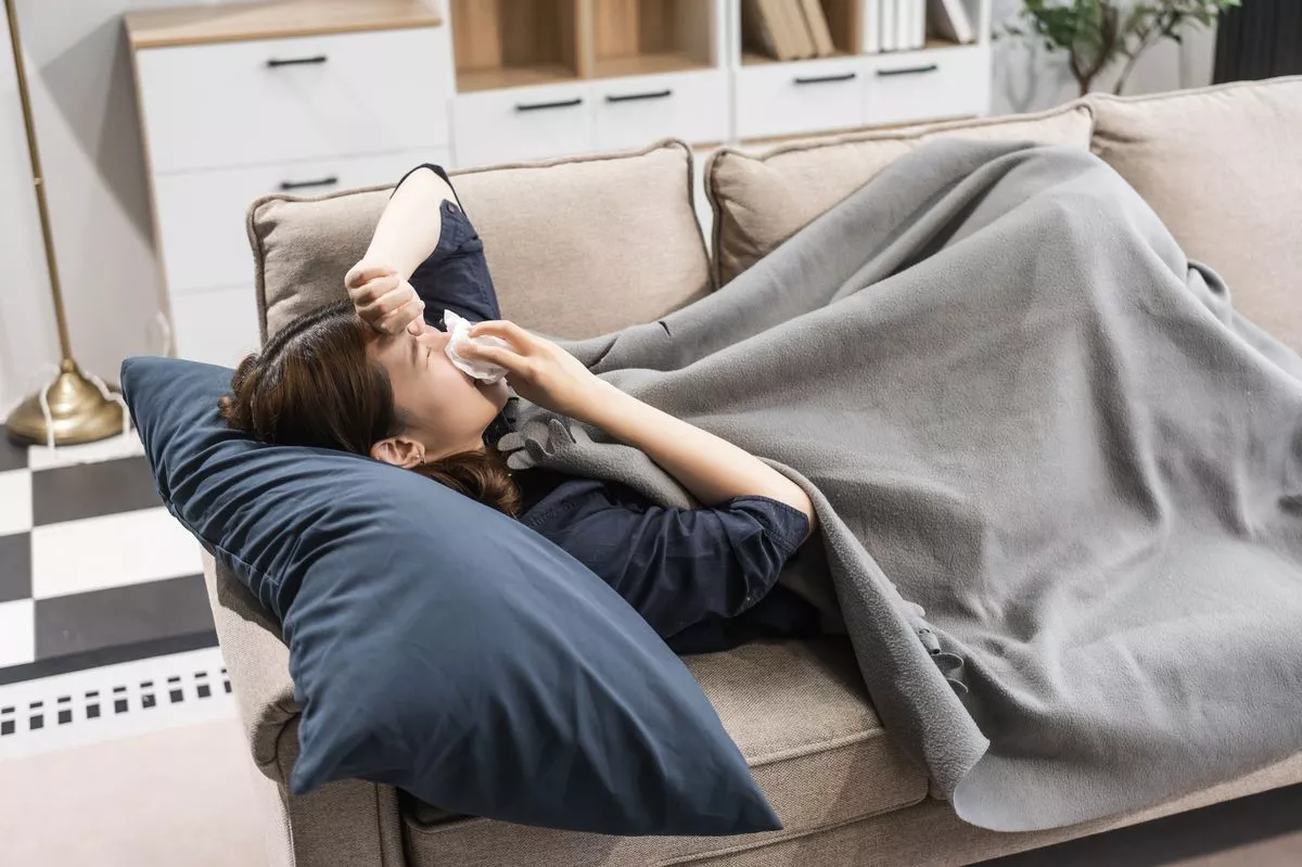 A young woman resting on a sofa, suffering from the flu. She shows symptoms like fever, chills, body aches, and fatigue, requiring rest and care.