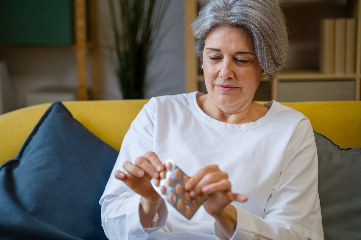 Mature woman extracting pills from blister pack, sitting on sofa in living room