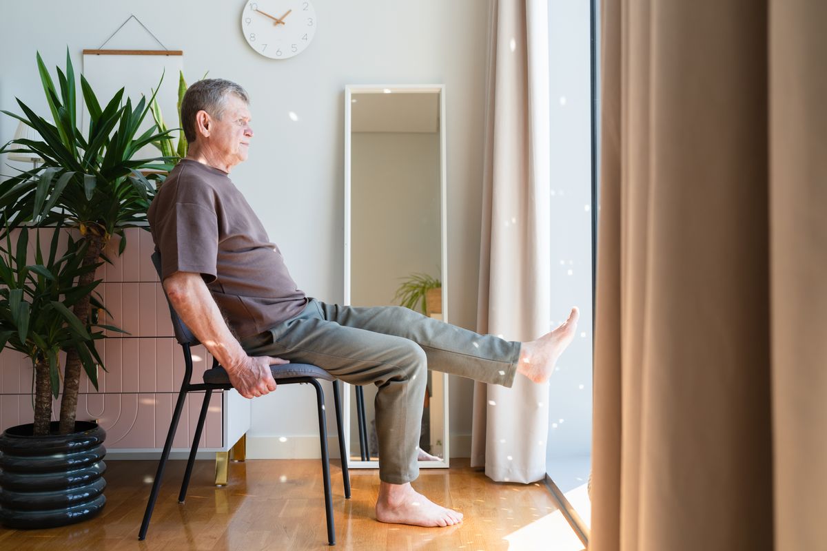 Senior man with grey hair is stretching at home while lifting up his leg