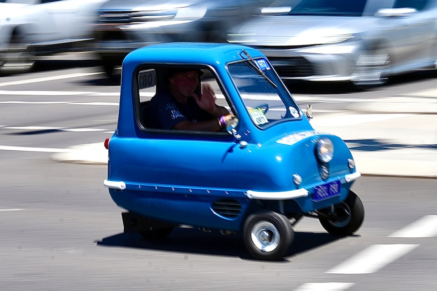 A vintage mini blue car drives down the road.