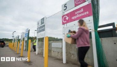 A woman carries a box as she crossed the car park of a recycling centre. A worker in a yellow high vis vest is seen entering the car park as well. The woman's figure is blurred. She is passing by a red sign that reads "Small appliances". A car with an open boot. It is a cloudy day.