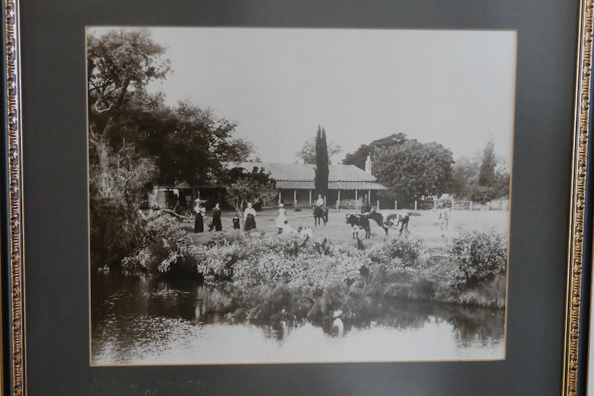 Black and white photo of people standing out the front of a house by a river