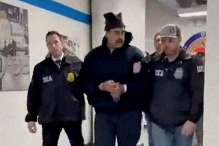 Nicolas Maduro, handcuffed and wearing jacket and beanie, flanked by US officials.