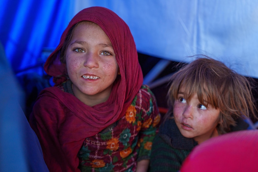 Two Afghan children sit in a tent, one smiles while the other looks on.