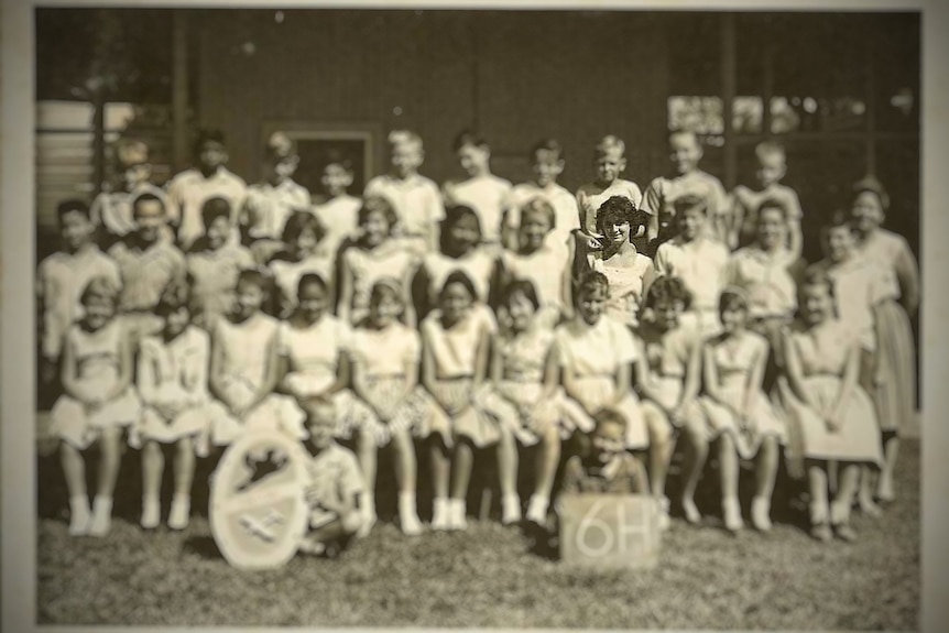 A sepia-toned, black and white, class photo filled with young girls and boys and a female teacher standing to the right.