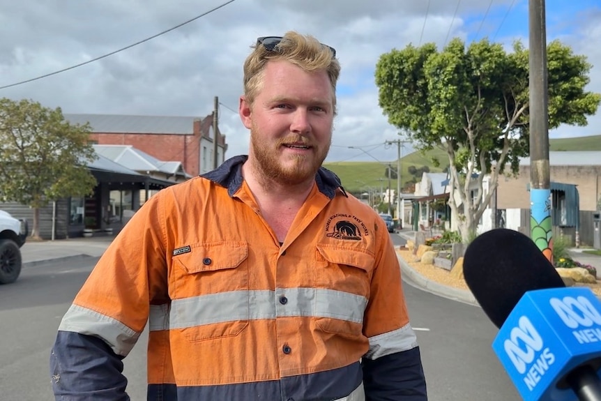 A photo of a man with blonde hair and a beard, wearing a orange fluorescent construction shirt, standing in a quiet street