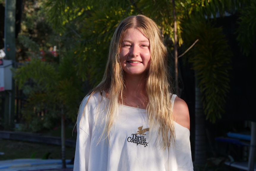 Girl with long blonde hair and an off the shoulder t-shirt, smiling directly at the camera with the sun shining on her face.