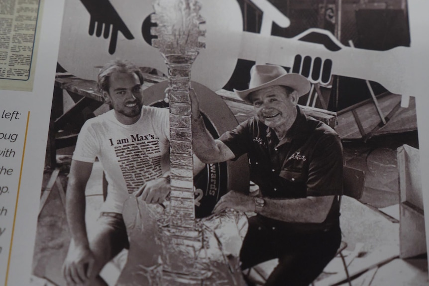 A black and white photo of two men hold up a giant golden guitar.