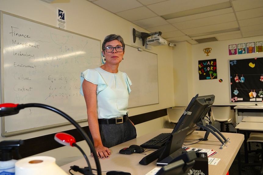 A woman stands behind a desk in a university classroom with a whiteboard behind her.