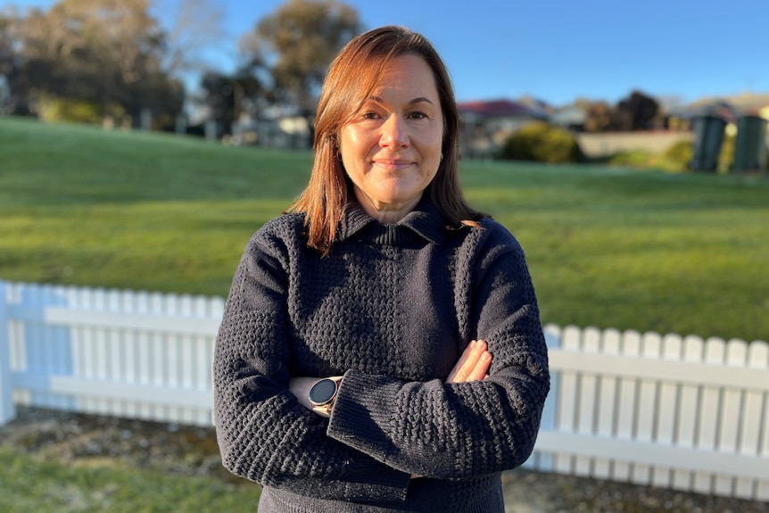A woman standing with her arms crossed in front of a low picket fence and a grassy hill
