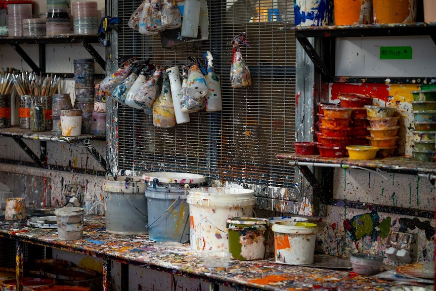 A bench stacked with buckets, small containers and spray bottles of paint, and jars of paint brushes.