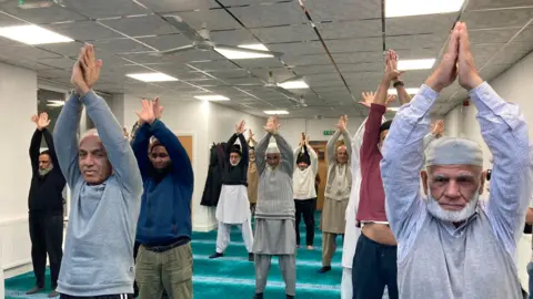 Cathy Minton/BBC A pilates class is held in a mosque in Bradford. The men participating are in comfortable, casual clothing and traditional dress. All are standing with their arms raised in the air.