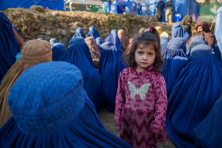A young Afghan girl stands with women in a camp.