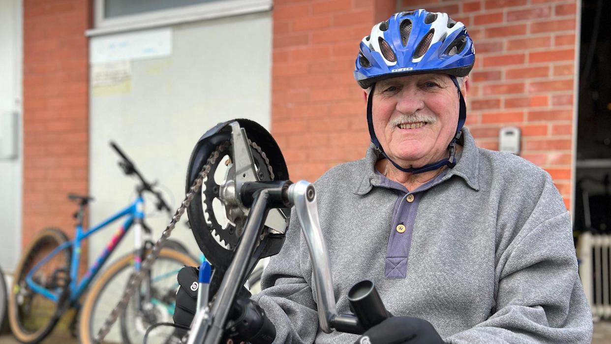 Alan is wearing a helmet and gloves, adjusting the handlebars of a red adapted tricycle outside a brick building, with several bicycles parked nearby