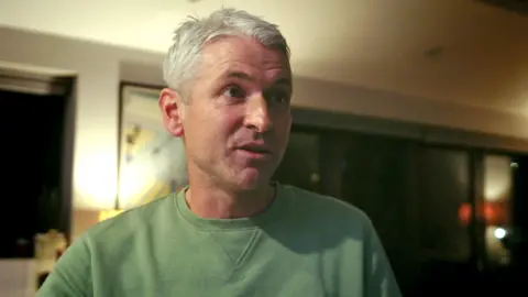 Rob Jones, with short grey hair wearing a green sweatshirt, stands indoors in a dimly lit room. The background features dark windows, a lit lamp, and framed artwork.