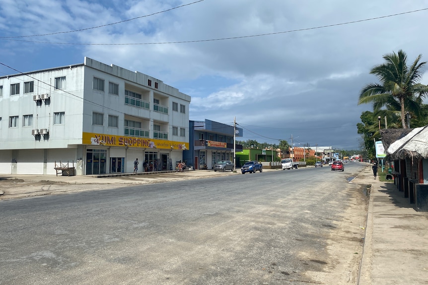 A wide dusty tar sealed street with some cars parked at the kerb and a car moving into the distance.