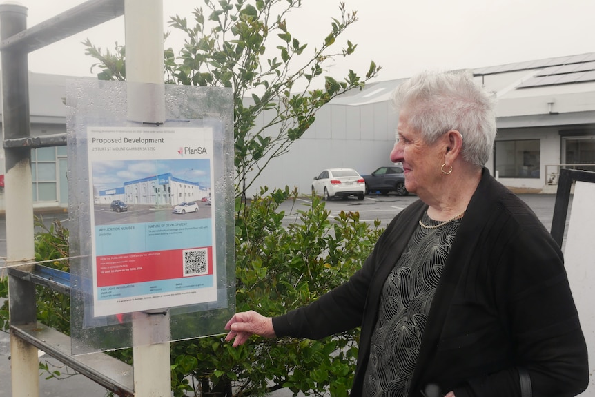 A grey-haired older woman is looking at a blue and red laminated paper sign outside a carpark on a rainy day.