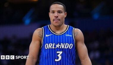 Desmond Bane seen from waist up in the centre of the image looks on in a blue Orlando Magic vest with white vertical pinstripes. A white number three is in the centre of his chest and above it the word 'Orlando' in white, with the letter A replaced by a grey five-pointed star