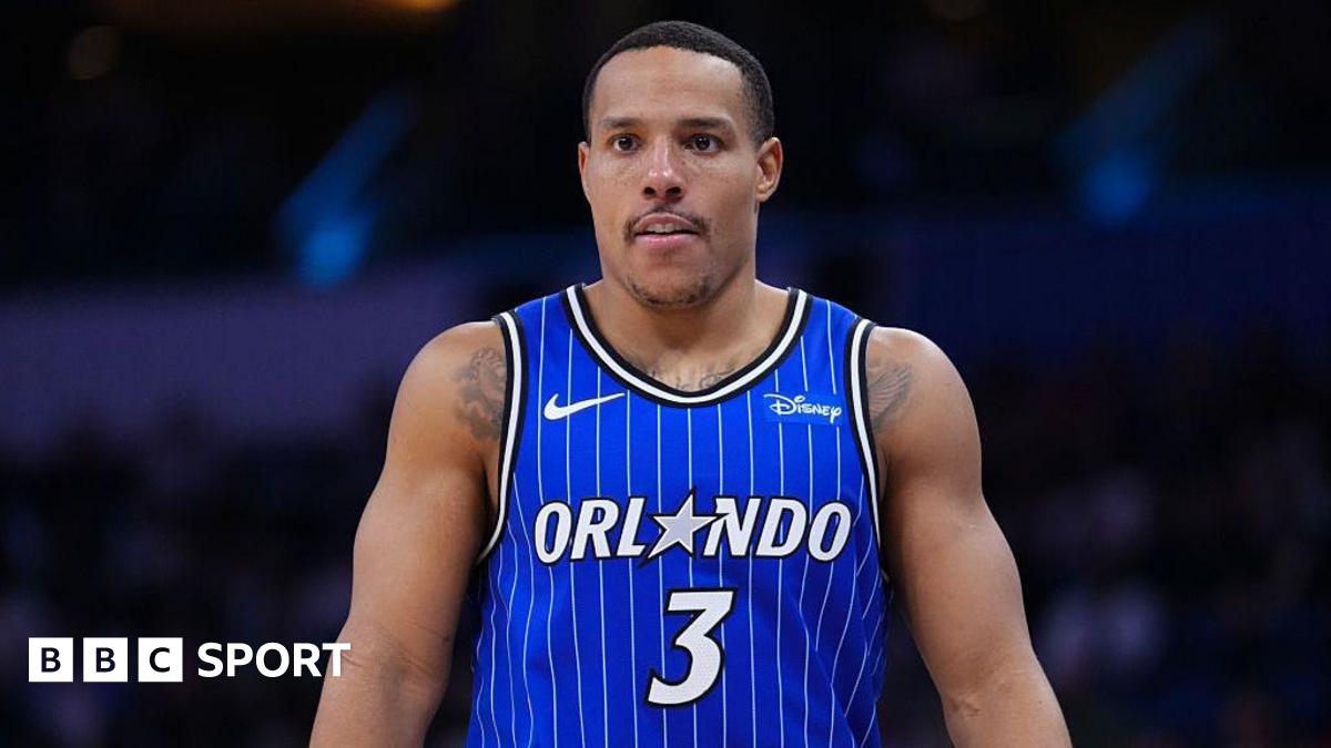 Desmond Bane seen from waist up in the centre of the image looks on in a blue Orlando Magic vest with white vertical pinstripes. A white number three is in the centre of his chest and above it the word 'Orlando' in white, with the letter A replaced by a grey five-pointed star
