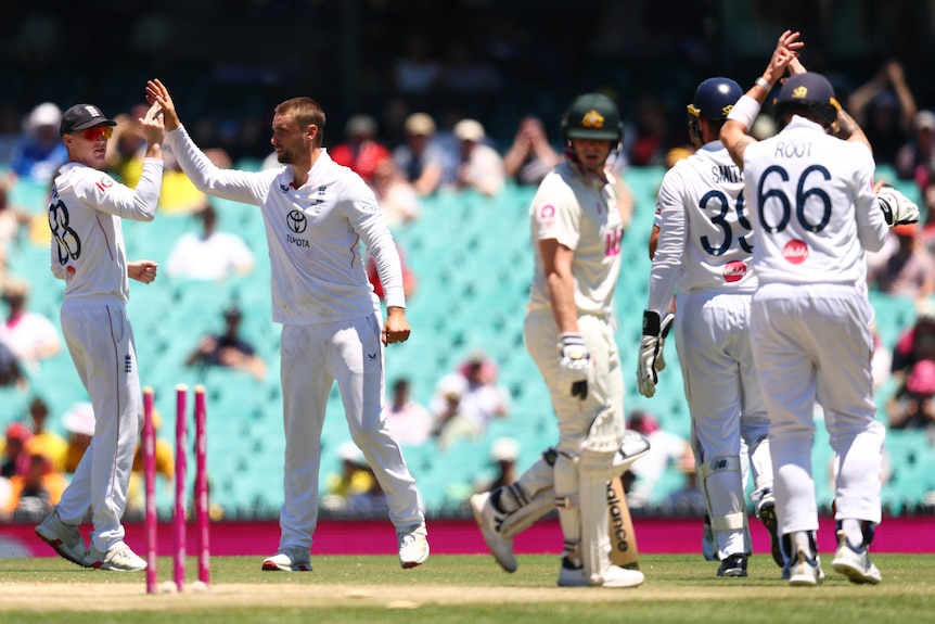 A cricket batter looks behind him as the stumps have the bails off and other players celebrate
