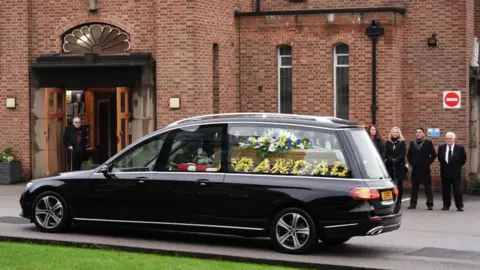PA Media Floral tributes in and around a coffin carried in a hearse, including one saying 'Grandad' and another with white, yellow and blue flowers. A group of people are lined up outside the church.