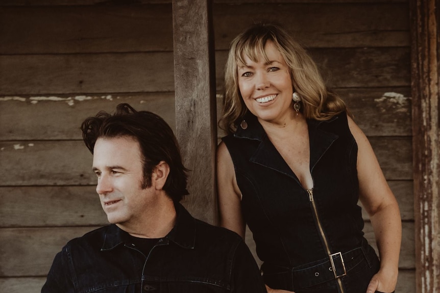 A man and a woman, both dressed in black, stand side by side outside an old wooden house.