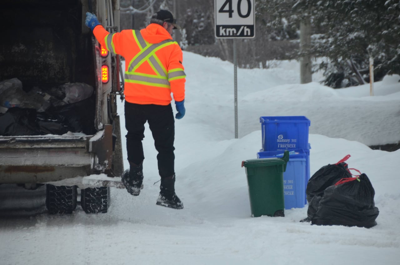 A man in an orange vest hops off a truck and moves toward recycling bins and garbage bags