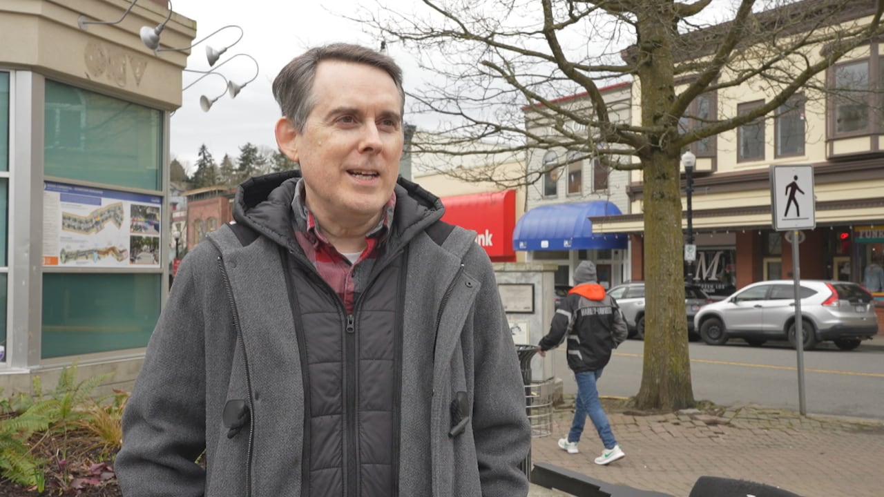 A man with short grey hair stands outdoors in a grey jacket. 