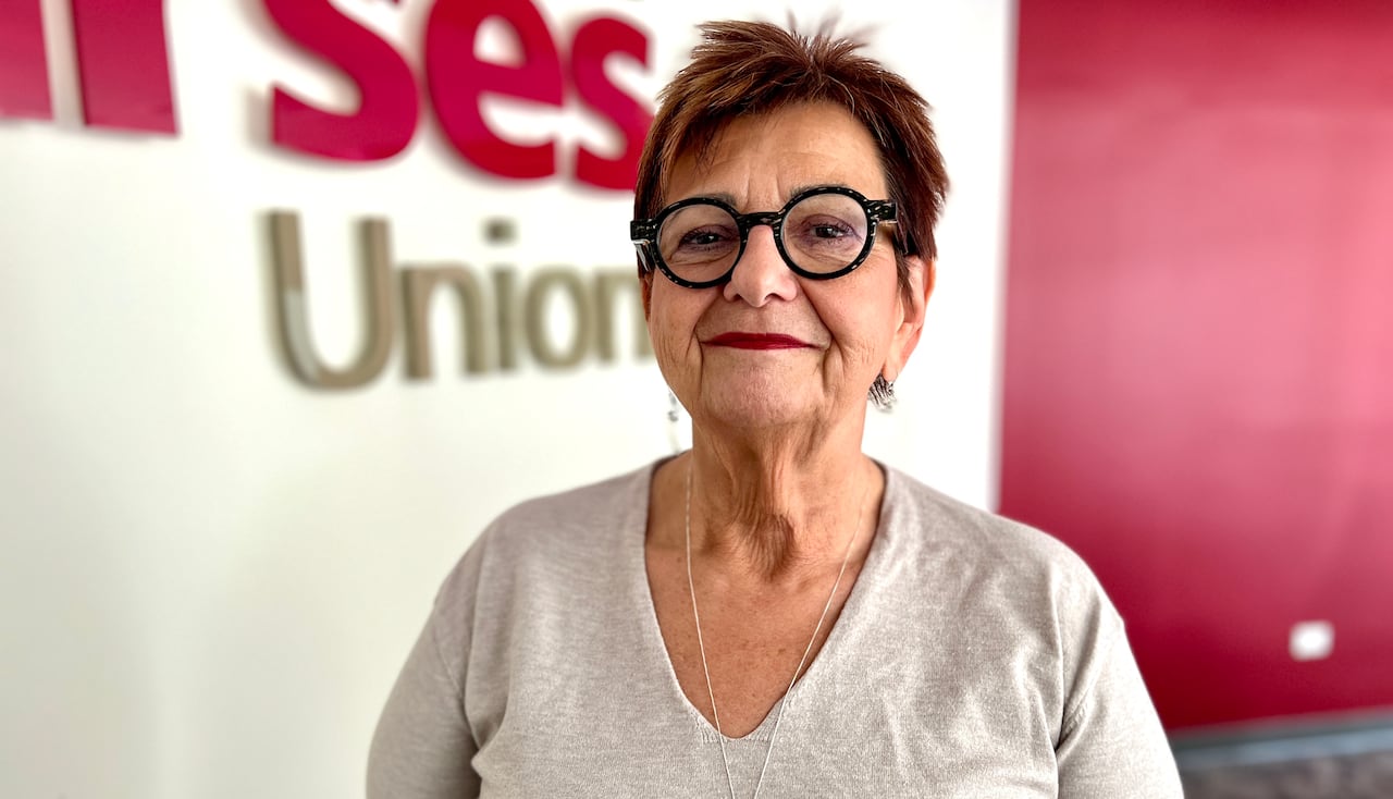 A woman with short hair and wearing a grey v-neck shirt stands and smiles. Behind her is a sign that says, 'Manitoba Nurses Union.'