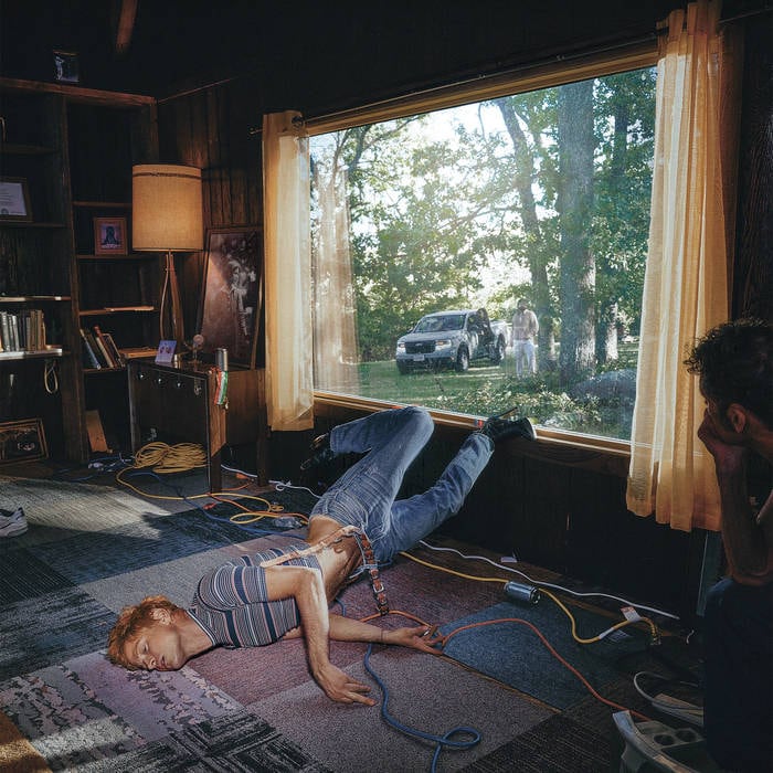 A person lays splayed out on a patchwork carpet, their feet against a window.