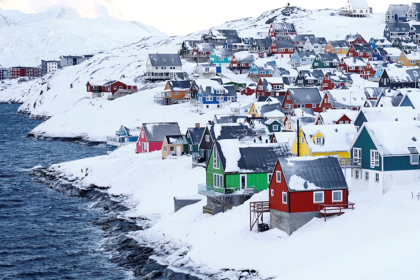 Green, red and yellow houses seen on the side of a white, snowy mountain alongside dark blue oceanwater