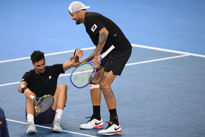 Two male tennis players dressed in black, one helping the other up from the ground on the court.