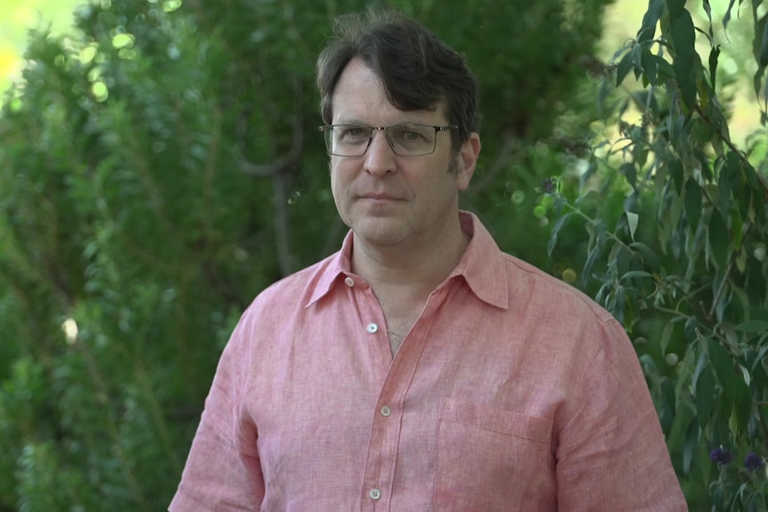 Daniel Aghion has dark hair and wears glasses and a pink collared shirt and stands in front of green bushes.