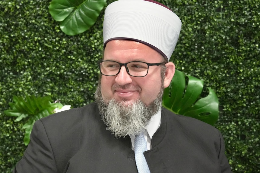 An imam smiles against a green, leafy backdrop.