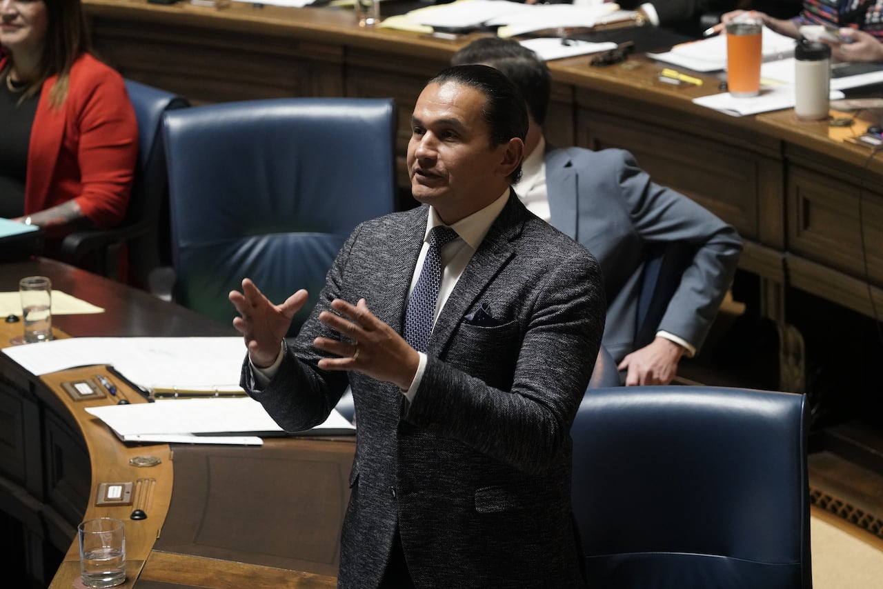A man in a dark suit gestures with his hands while speaking during a government legislative building.